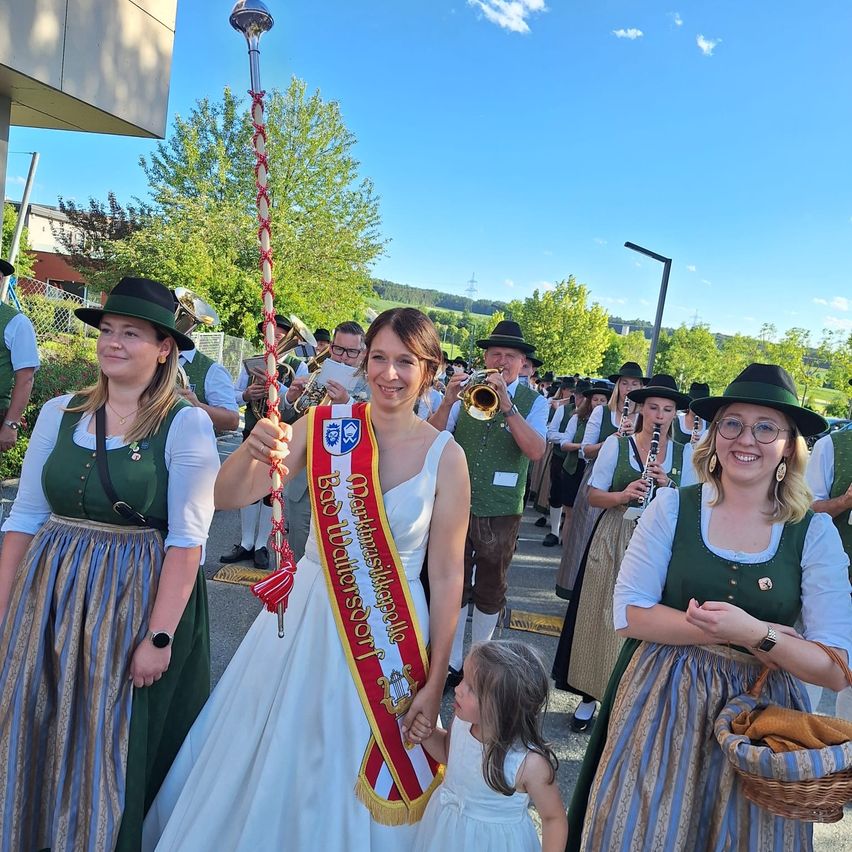 Eine Frau in einem weißen Kleid hält eine Schleife und einen Stab und steht mit einer Band hinter ihr. Sie sind in einem Umzug, mit mehreren Leuten in traditioneller deutscher Kleidung. Bäume und ein Gebäude sind im Hintergrund.