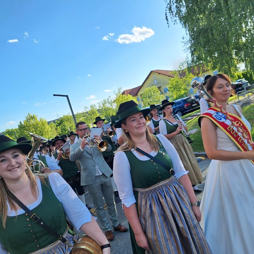 Eine Blaskapelle spielt Instrumente, während sie in traditioneller bayerischer Kleidung die Straße entlanggeht. Im Hintergrund steht ein Gebäude mit einem geparkten Auto.