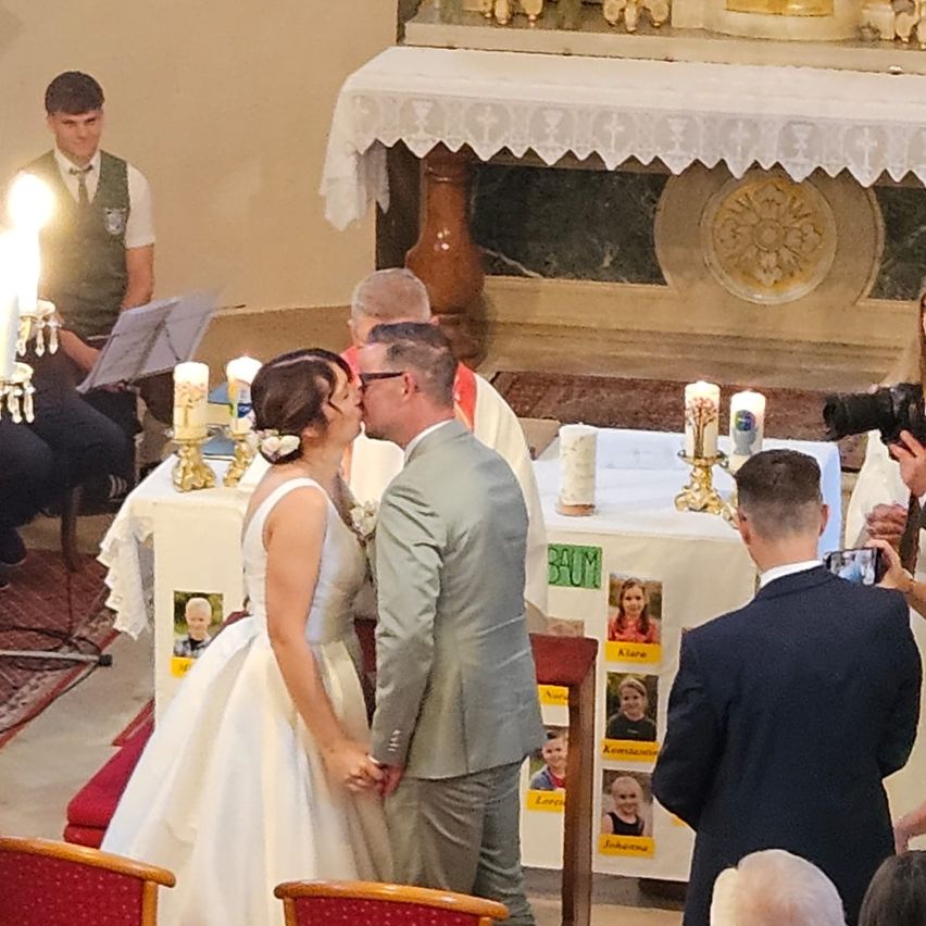 Ein frisch verheiratetes Paar küsst sich vor einem Altar. Sie sind von Gästen, Kerzen und einem Fotografen umgeben. Ein Priester und ein Musiker sind ebenfalls anwesend.