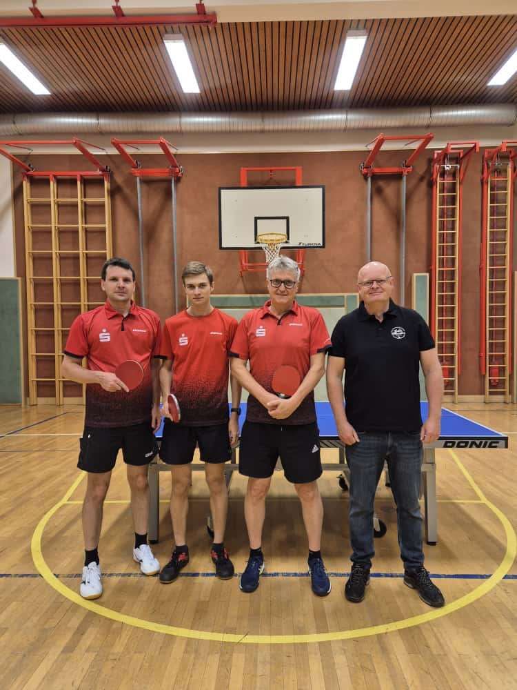 Vier Männer stehen in einer Turnhalle. Sie tragen rote und schwarze Sporttrikots. Drei von ihnen halten Schläger. Ein Tischtennistisch steht vor ihnen. Dahinter befinden sich rote Leitern und ein Basketballkorb.