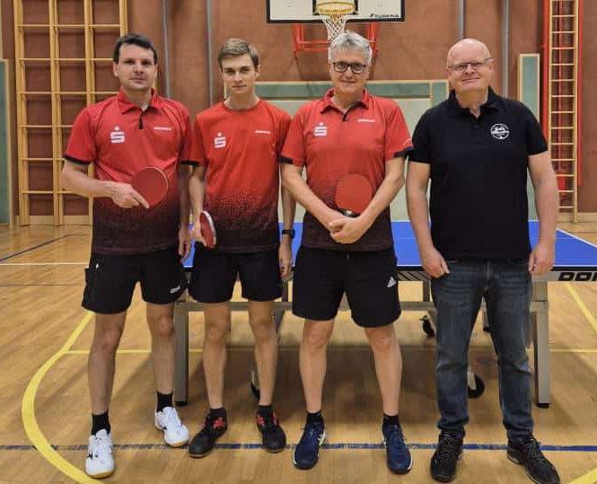 Vier Männer stehen vor einem Tischtennistisch, bekleidet mit Sportkleidung. Zwei halten Schläger, und zwei haben die Hände in den Taschen. Der Ort ist eine Indoor-Sportanlage.