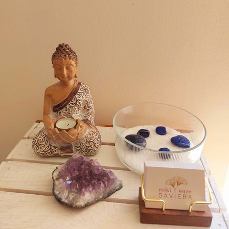 Ein Holztisch zeigt eine Buddha-Figur mit einer Kerze, eine Schale mit Steinen und eine Visitenkarte mit 'Reiki Oasis Saviera.'