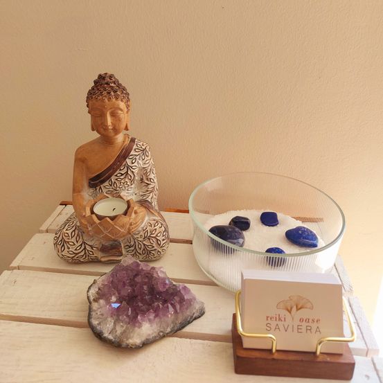 Ein Holztisch zeigt eine Buddha-Figur mit einer Kerze, eine Schale mit Steinen und eine Visitenkarte mit 'Reiki Oasis Saviera.'