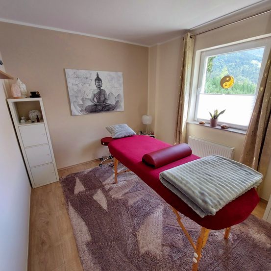 Ein Massageraum mit einem roten Massagetisch, Kissen und einem Teppich. Ein weißer Schrank, ein Gemälde und ein Fenster sind im Raum. Das Fenster zeigt einen Blick auf Berge.