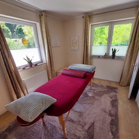 Ein Massagetisch mit zwei Kissen und zwei gerollten Handtüchern steht in einem Raum mit zwei Fenstern. Auf der Fensterbank stehen zwei Topfpflanzen und ein Heizkörper an der Wand.