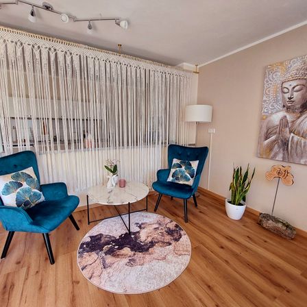 Ein gemütliches Wohnzimmer mit blauen Stühlen und einem runden Tisch, einer Makramee-Gardine, einem Buddha-Gemälde, einer Lampe und einer Topfpflanze.