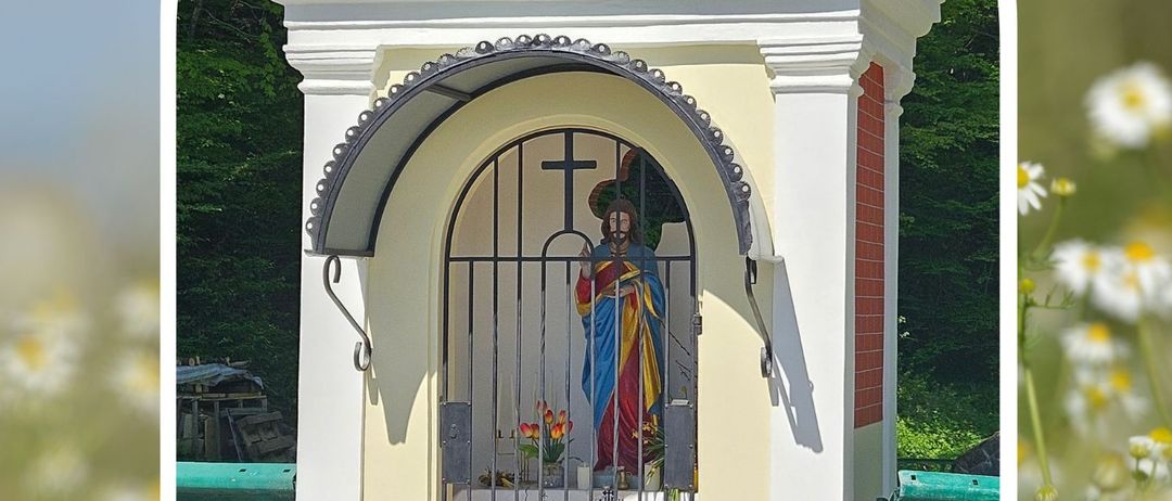 Eine kleine Kapelle mit einer Statue im Inneren, umgeben von Blumen und Grün. Die Kapelle hat ein Kreuz und das Jahr 1854. Sie ist renoviert und für den Gottesdienst geöffnet.