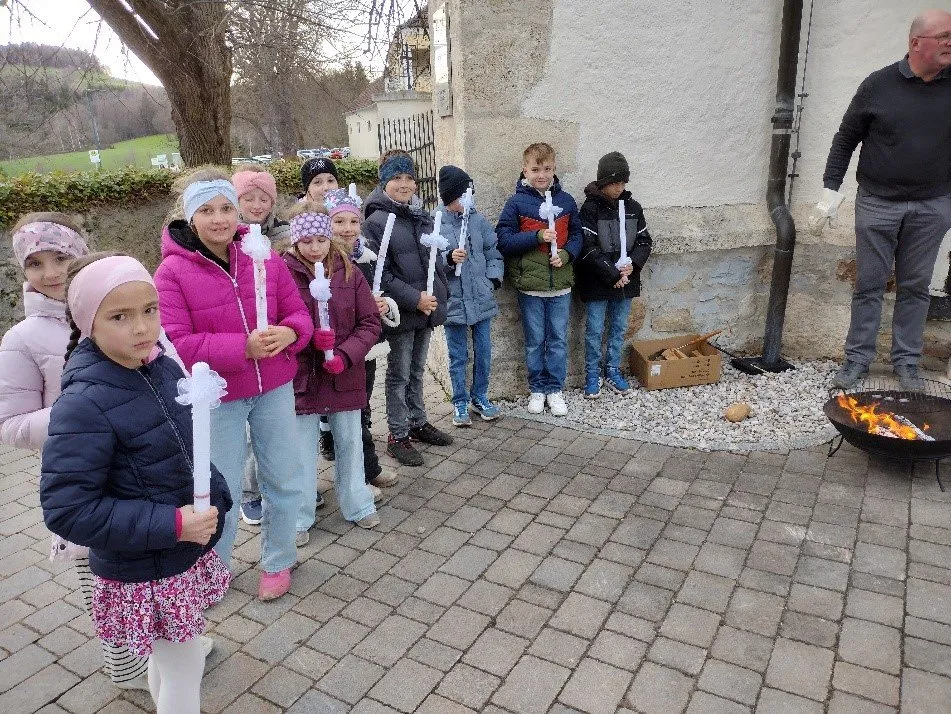 Eine Gruppe von Kindern mit weißen Kerzen steht in einer Reihe bei kaltem Wetter. Dahinter befindet sich eine Wand, und ein Feuerkorb ist in der Nähe.