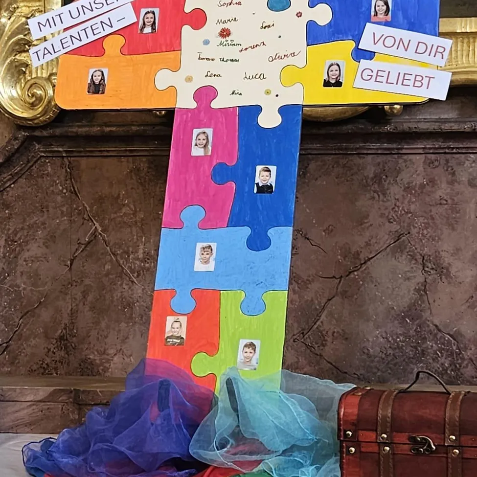 Ein farbenfrohes Kreuz aus Puzzleteilen mit Bildern von Kindern und den Worten 'Mit unseren Talenten' und 'Von dir geliebt' darauf. Es steht auf einer Marmoroberfläche mit einem Koffer in der Nähe.