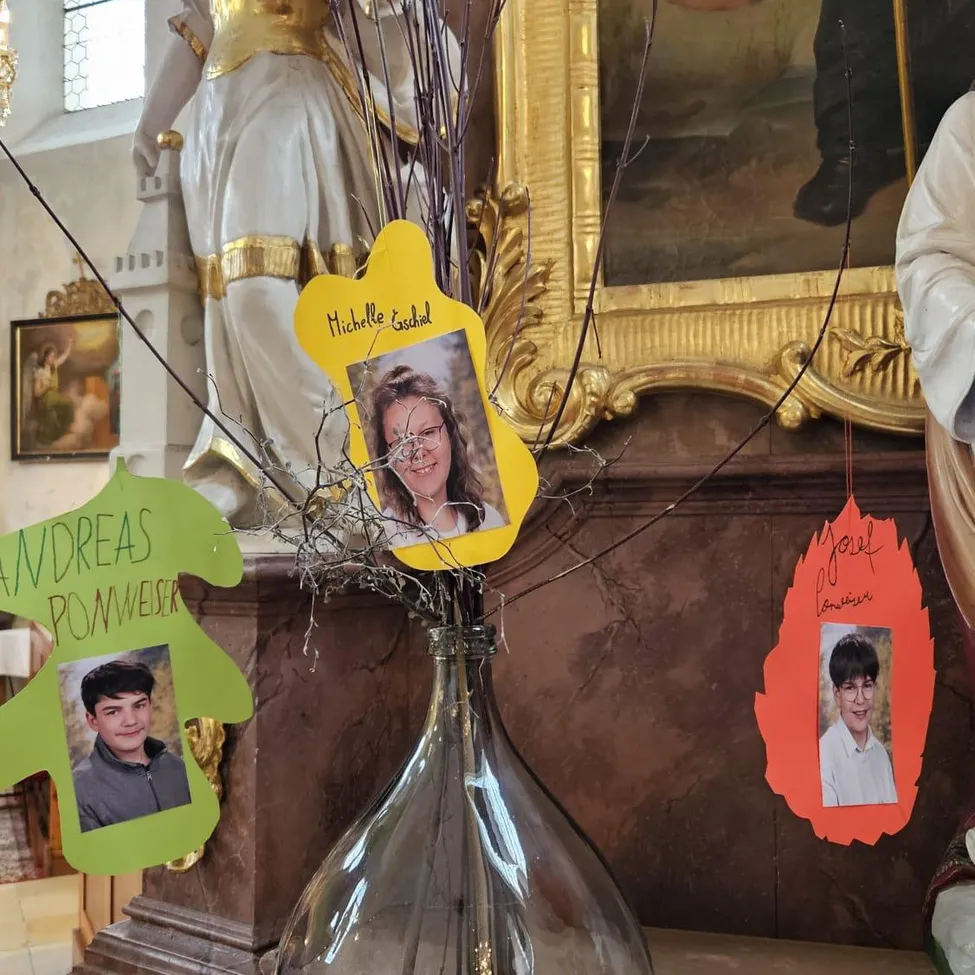 Vor einem goldenen Bilderrahmen steht eine Glasvase mit drei Fotokarten. Die Vase hat grüne, gelbe und orange Karten. Die Fotokarten tragen die Namen Michelle, Andreas und Joel.