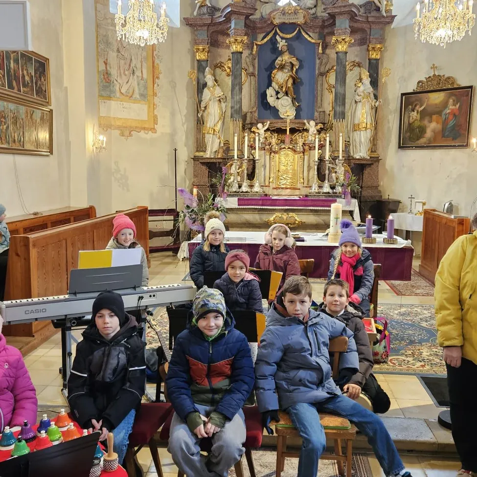 Eine Gruppe von Kindern sitzt in einer Kirche, einige in Winterkleidung. Vor ihnen steht ein Klavier mit einer Glockenreihe. Dahinter befindet sich ein Altar mit einer Statue und Kerzen.