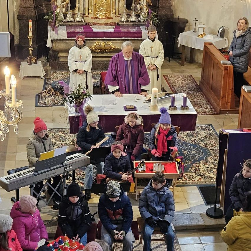 In einer Kirche spielen Kinder während eines Gottesdienstes Musikinstrumente. Ein Priester steht hinter einem Tisch und liest aus einem Buch. Der Raum ist mit Kerzen geschmückt, und Menschen sitzen herum.