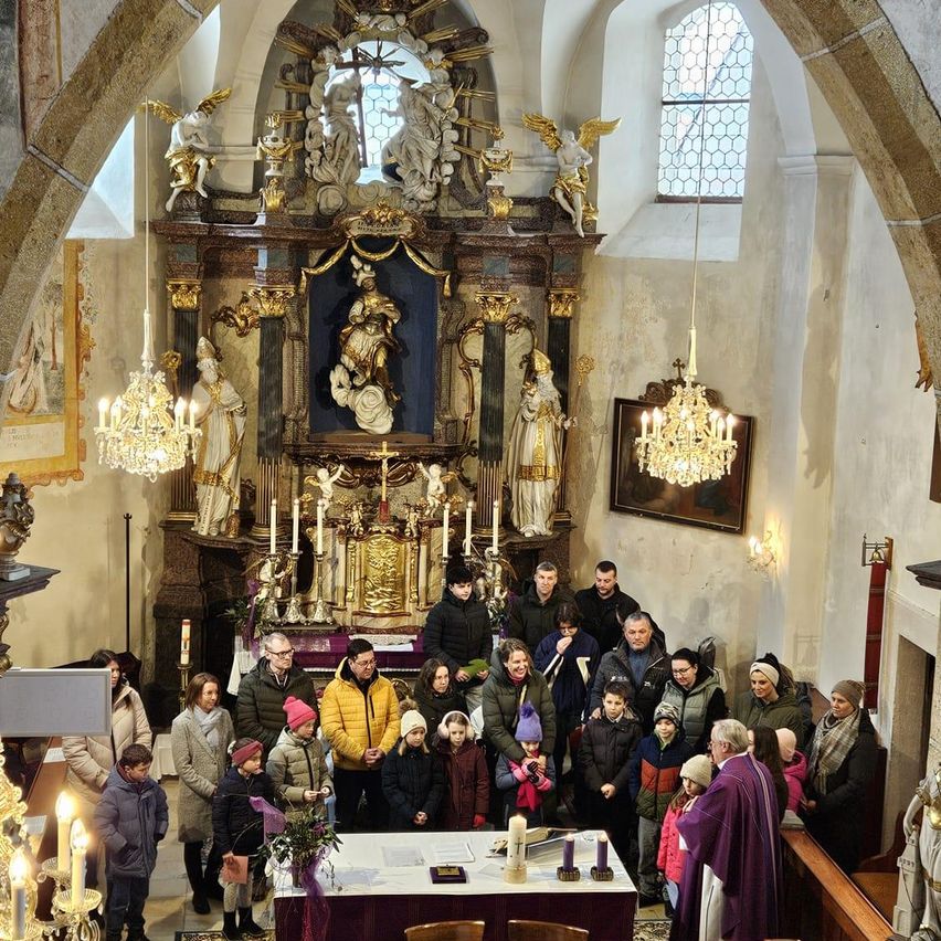 Eine Gruppe von Menschen in Winterkleidung steht in einer Kirche und schaut zu einem Priester. Der Altar ist mit Kerzen und Blumen geschmückt, und die Kirche verfügt über kunstvolle Statuen und Kronleuchter.