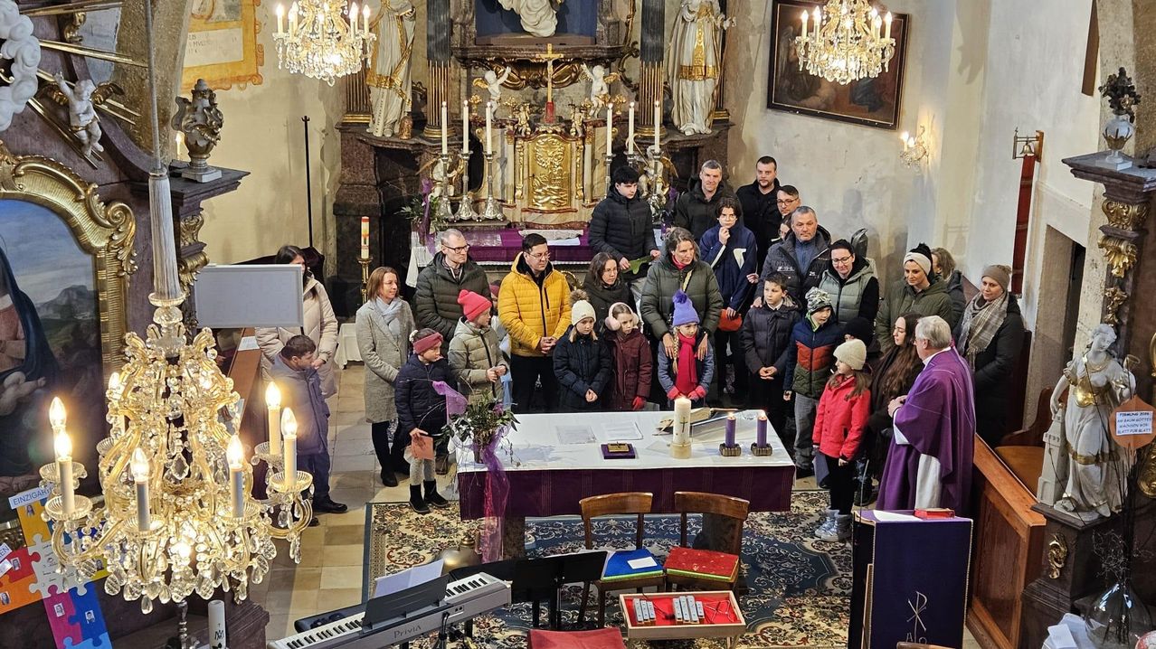 Eine Gruppe von Erwachsenen und Kindern steht um einen Altar in einer Kirche, Kerzen und religiöse Gegenstände sind vorhanden.