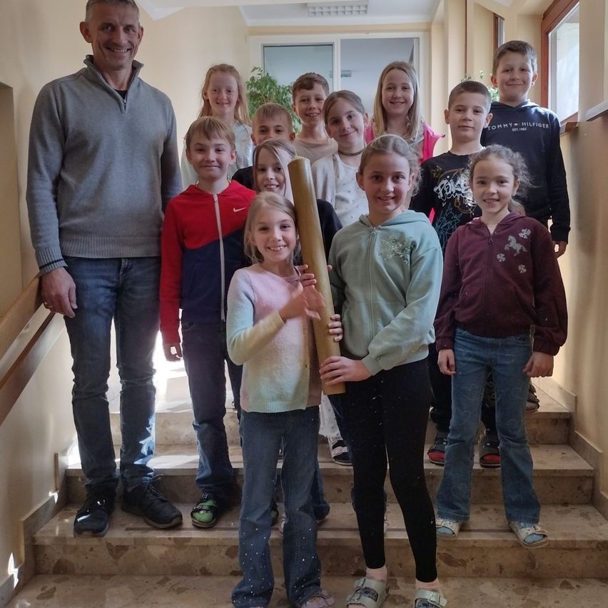 Eine Gruppe von Kindern und zwei Erwachsenen posiert für ein Foto auf einer Treppe, ein Mädchen hält ein großes Objekt, alle tragen Freizeitkleidung.