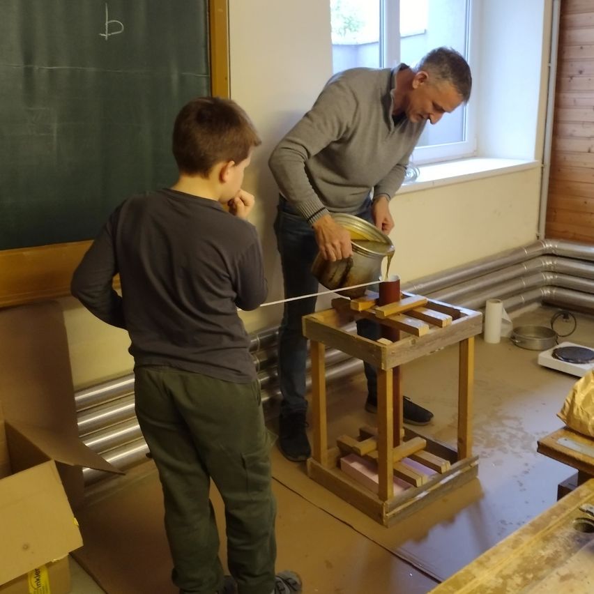 Ein erwachsener Mann und ein Junge befinden sich in einem Raum mit einer Tafel. Der Mann gießt Flüssigkeit in einen Behälter auf einem Holzständer.
