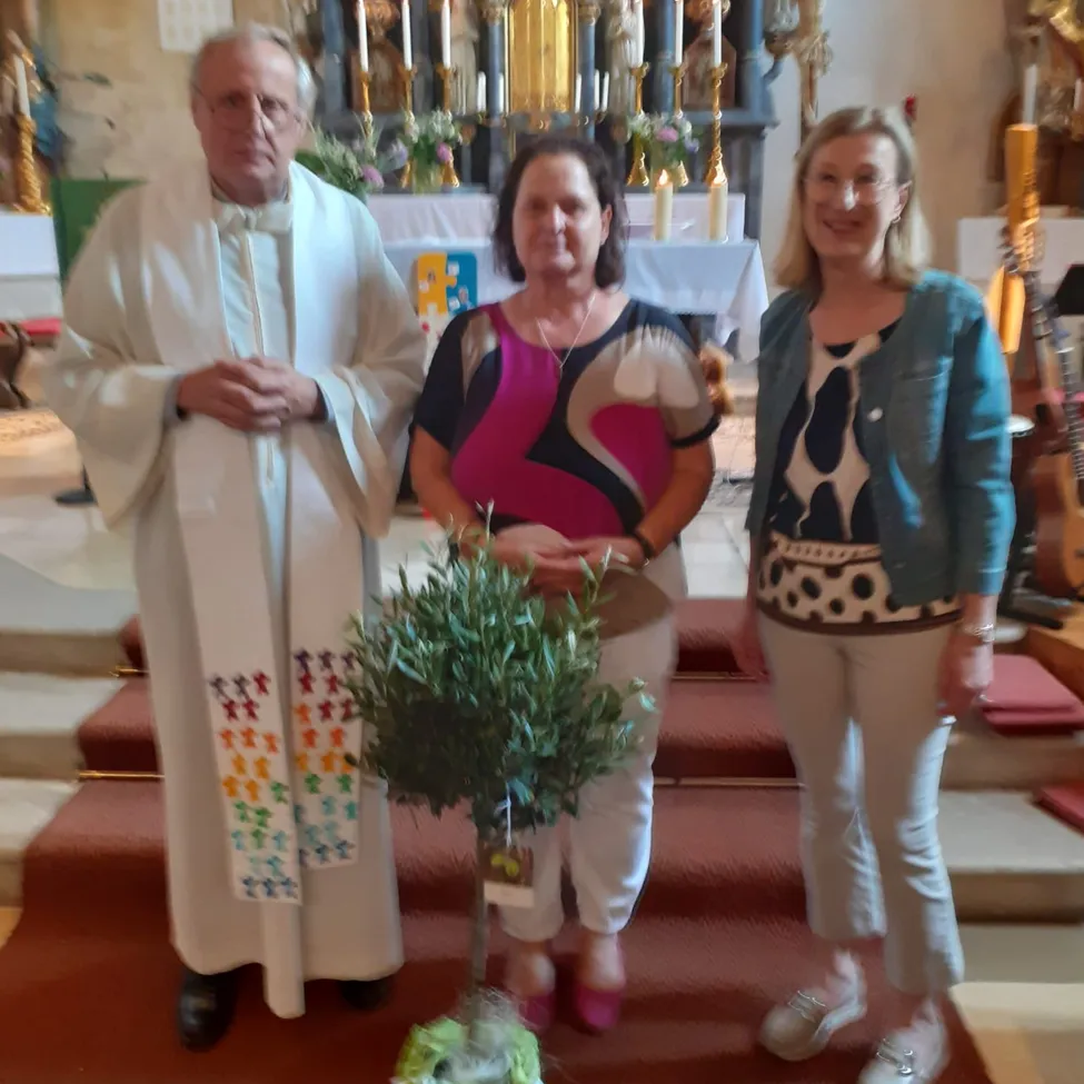 Drei Personen stehen in einer Kirche, ein Priester mit Brille und zwei Frauen. Eine hält einen Topf mit einem Olivenbaum. Hinter ihnen steht ein Altar mit Kerzen und eine Gitarre auf der rechten Seite.