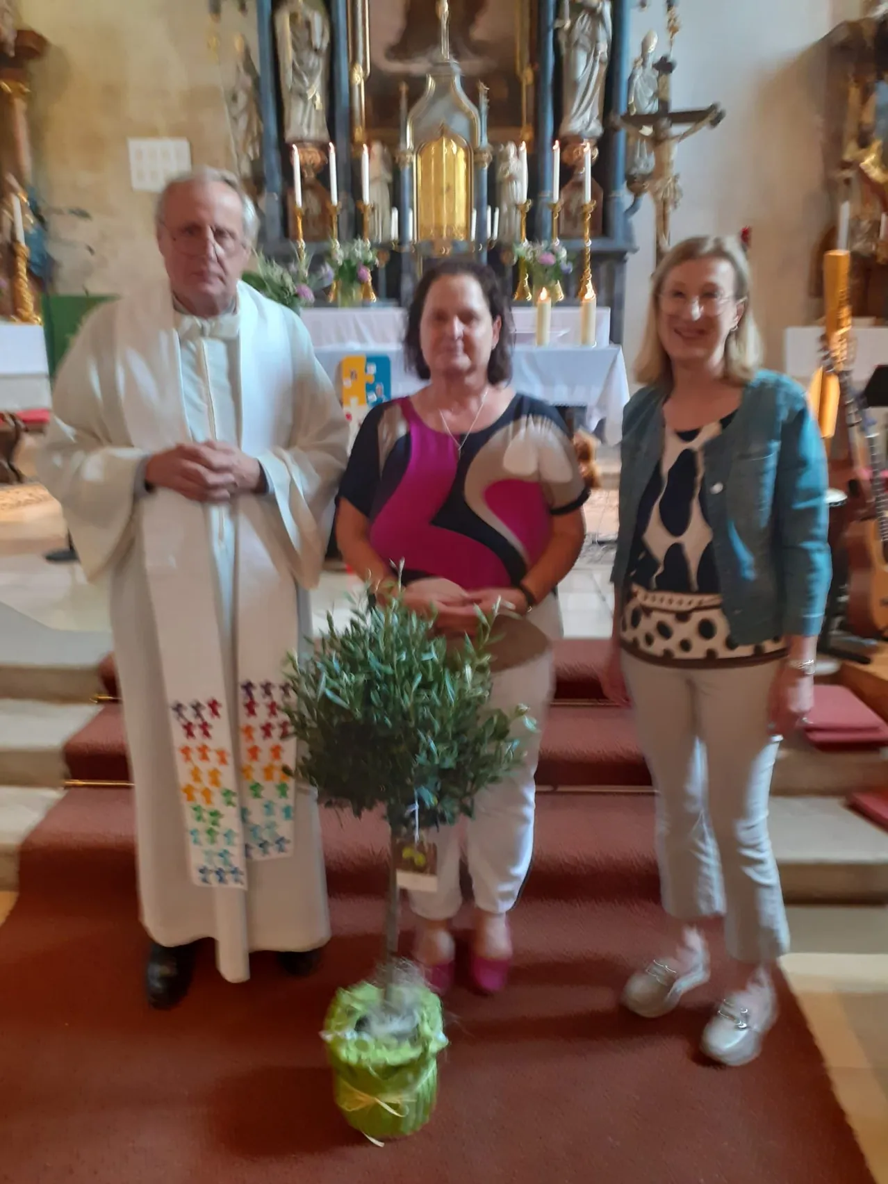 Drei Personen stehen in einer Kirche, ein Priester mit Brille und zwei Frauen. Eine hält einen Topf mit einem Olivenbaum. Hinter ihnen steht ein Altar mit Kerzen und eine Gitarre auf der rechten Seite.