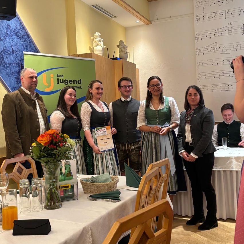A group of people are standing around a table with a flower arrangement, posing for a photo. They are wearing traditional German attire. Behind them, a wall displays musical notes. A banner is mounted on the wall, and a desk with a glass and a cell phone is visible.