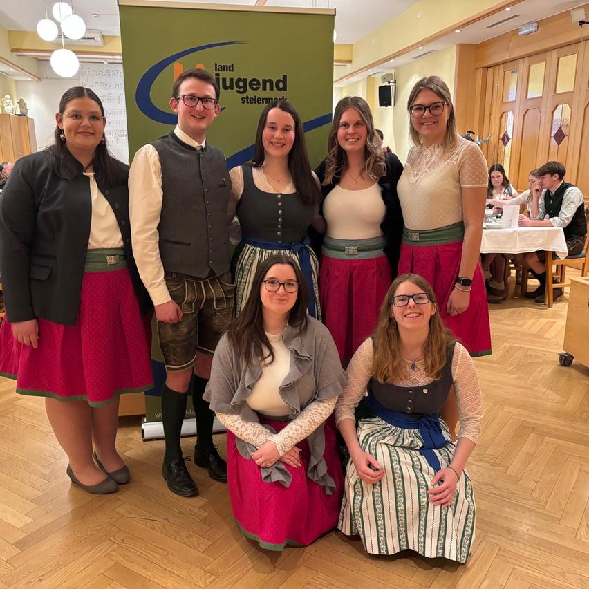 A group of six people, three women and three men, are posing for a photo in a room, possibly at an event. They are all wearing traditional German attire. The woman on the right is wearing glasses and a necklace. Behind them is a banner with the text 'landjugend steilmark'. In the distance, there are people sitting on chairs, possibly eating.