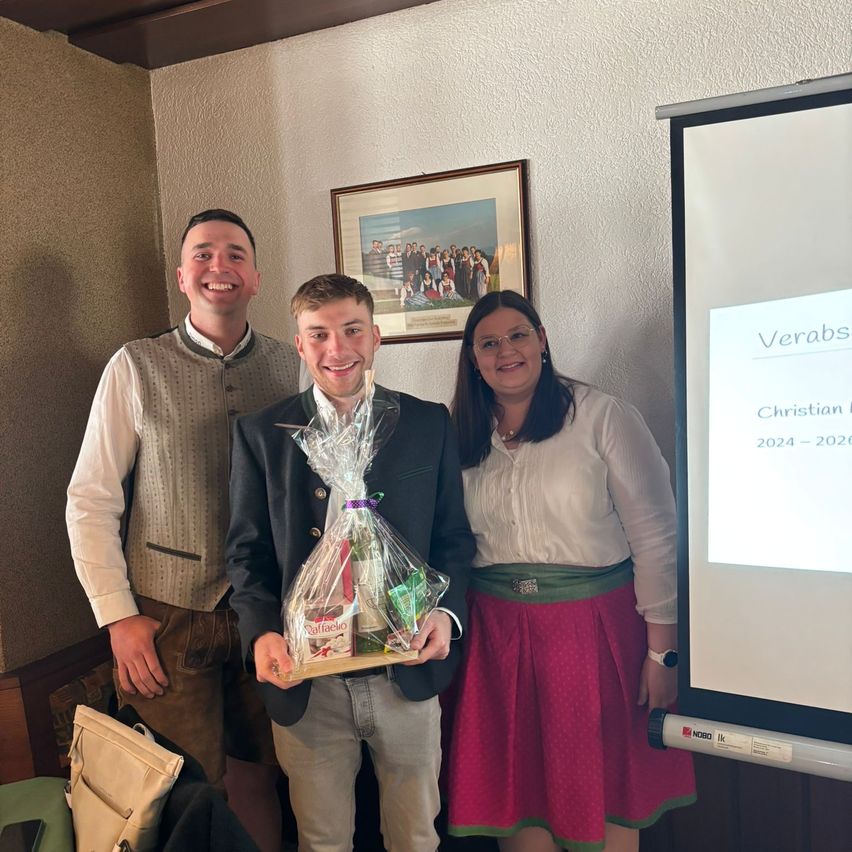 Three individuals are standing in a room, smiling. The man in the middle is holding a gift basket. The woman on the right is wearing a watch. Behind them is a projector screen with the words Verabs Christian 2024-2026.