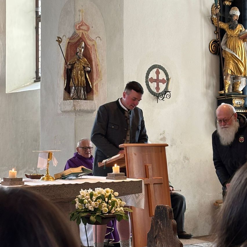 A man stands at a pulpit in a church, next to a priest in purple robes, while a man with a white beard kneels. The room features candles, flowers, and statues.
