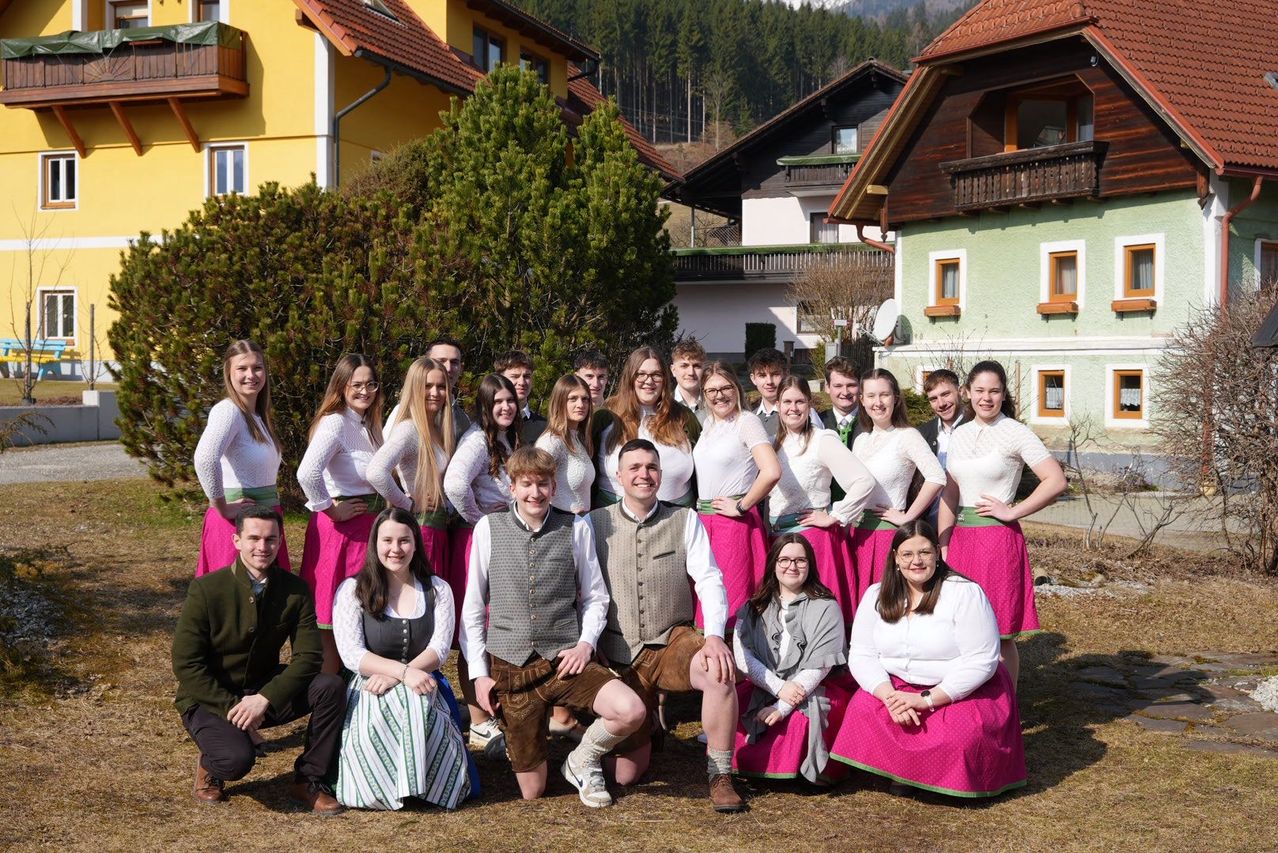 A group of people dressed in traditional attire are posing for a photo in front of houses. They are smiling and standing close to each other. Behind them are houses with balconies and trees.