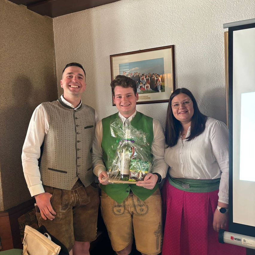 Three people dressed in traditional Bavarian clothing stand close together, smiling. The man in the middle holds a gift bag. They are in a room with a projector screen on the right and a framed picture on the wall behind them.