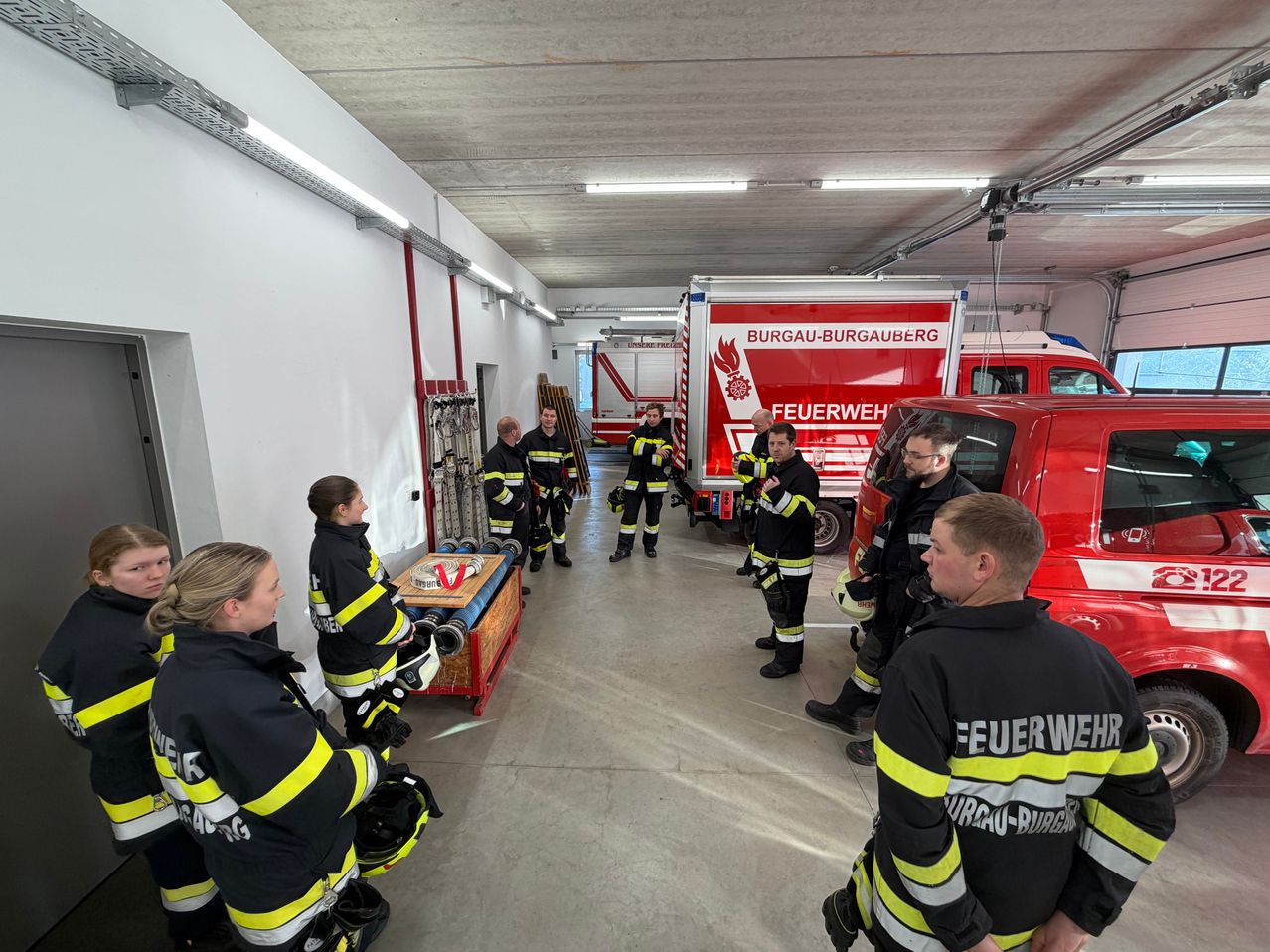 Feuerwehrleute sind in einer Station versammelt. Sie tragen Uniformen und Helme. Mehrere Feuerwehrwagen sind geparkt und Ausrüstung liegt auf dem Boden.