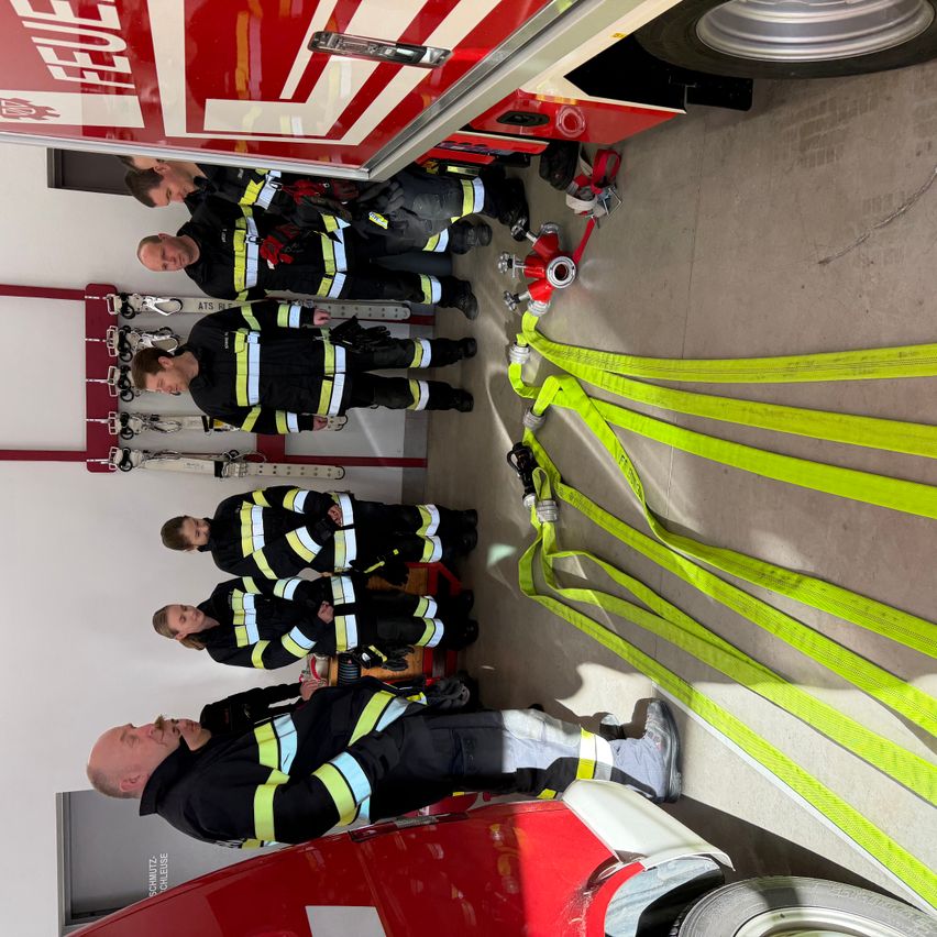 Eine Gruppe von Feuerwehrleuten befindet sich in einer Feuerwache, trägt Uniformen und steht in der Nähe eines Feuerwehrwagens, mit Sicherheitsausrüstung auf dem Boden.