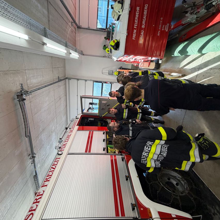 Mehrere Feuerwehrleute befinden sich in einer Feuerwache und bereiten einen Feuerwehrwagen vor. Ein Feuerwehrmann repariert die hintere Tür.