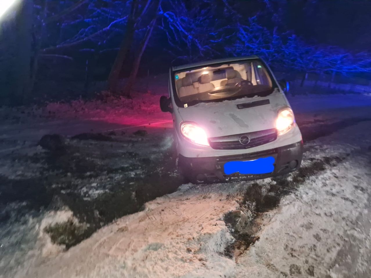 Ein weißer Van steckt im Schnee fest, mit eingeschalteten Scheinwerfern, umgeben von Bäumen und blauen Lichtern.