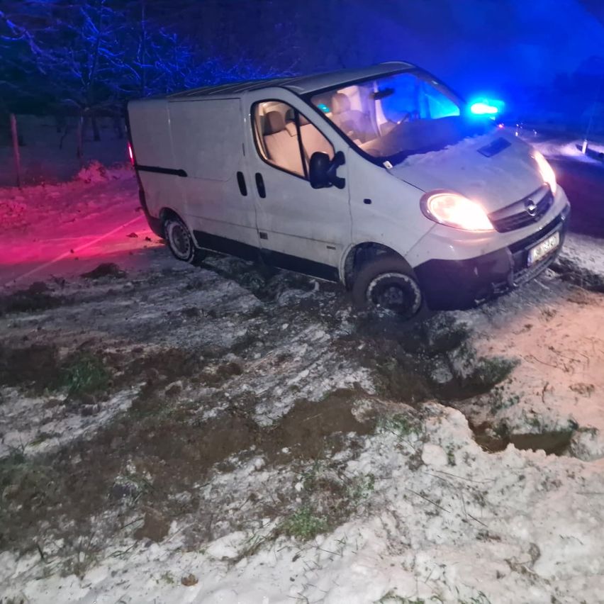 Ein weißer Lieferwagen steckt im Schnee fest, mit eingeschalteten Scheinwerfern, bei Nacht. Das Gebiet ist von blauen und roten Lichtern beleuchtet.