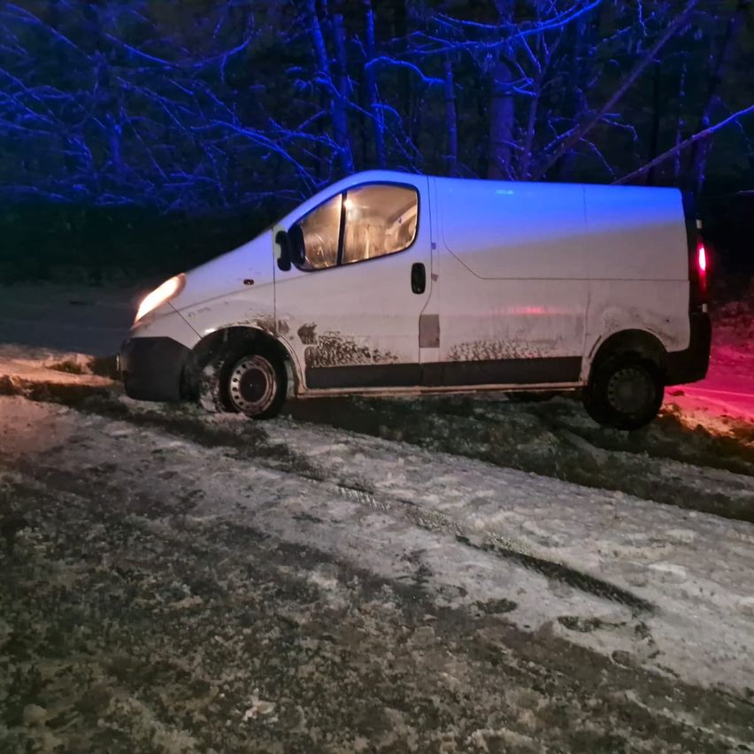 Ein weißer Lieferwagen steht an der Seite einer verschneiten Straße, ein Rad im Schnee. Bäume mit blauen Lichtern beleuchten den Hintergrund.