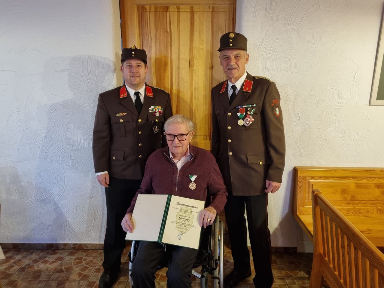 Ein älterer Mann im Rollstuhl hält ein offenes Zertifikat, während zwei Männer in Militäruniformen hinter ihm stehen. Einer der Männer hält eine Medaille und beide tragen Mützen.