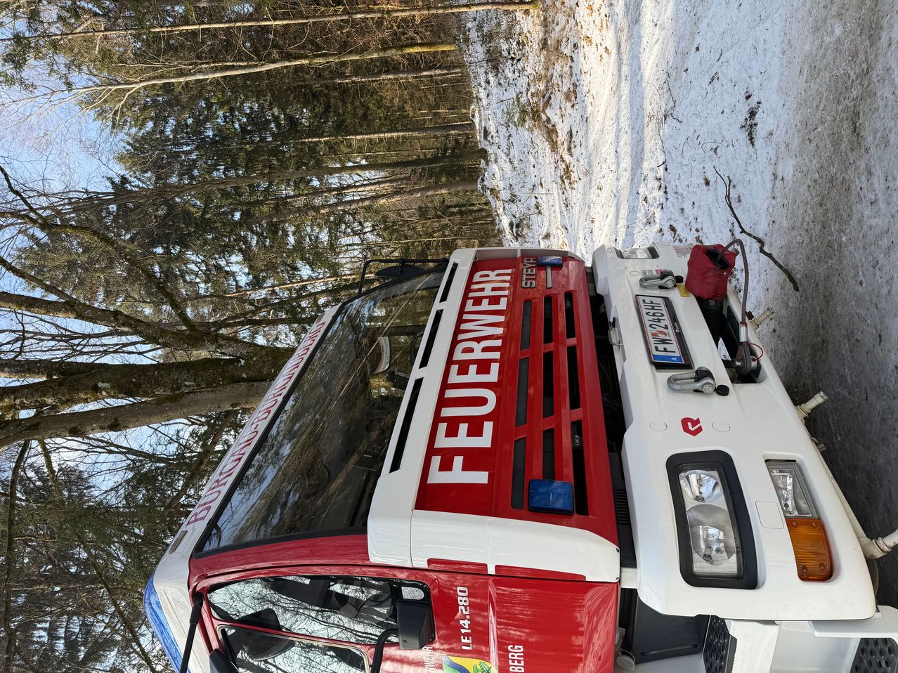 Ein Feuerwehrauto ist im Schnee geparkt, mit Bäumen und einem Berg im Hintergrund. Das Auto hat einen rot-weißen Körper und das Wort 'Feuerwehr' auf der Vorderseite geschrieben.