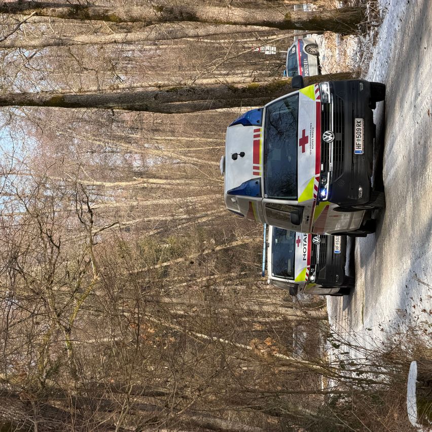 Ein Rettungswagen ist an der Seite einer verschneiten Straße geparkt, mit einem weiteren Fahrzeug dahinter. Ein Polizeiauto ist in der Ferne zu sehen. Das Gebiet ist von Bäumen umgeben.