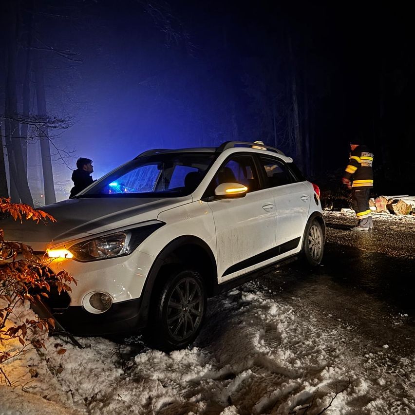Ein weißer SUV steckt bei Nacht im Schnee fest, mit einer Person daneben und einem Feuerwehrmann, der hilft.