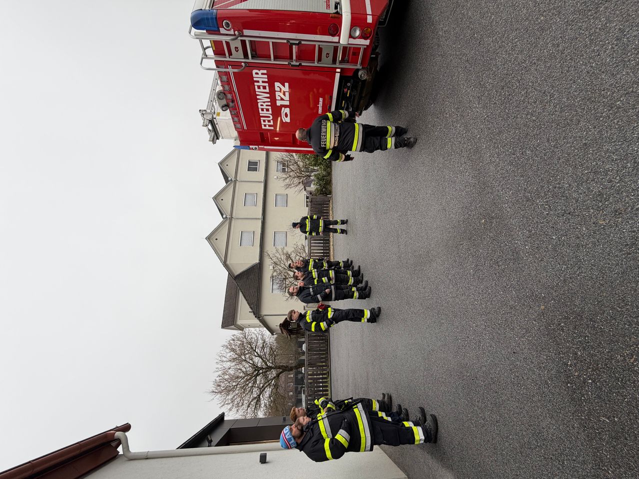 Eine Gruppe von Feuerwehrleuten steht in einer Reihe vor einem Gebäude. Das Gebäude hat viele Fenster. Die Feuerwehrleute tragen gelbe und schwarze Uniformen. Ein roter Feuerwehrwagen ist auf der Seite geparkt.