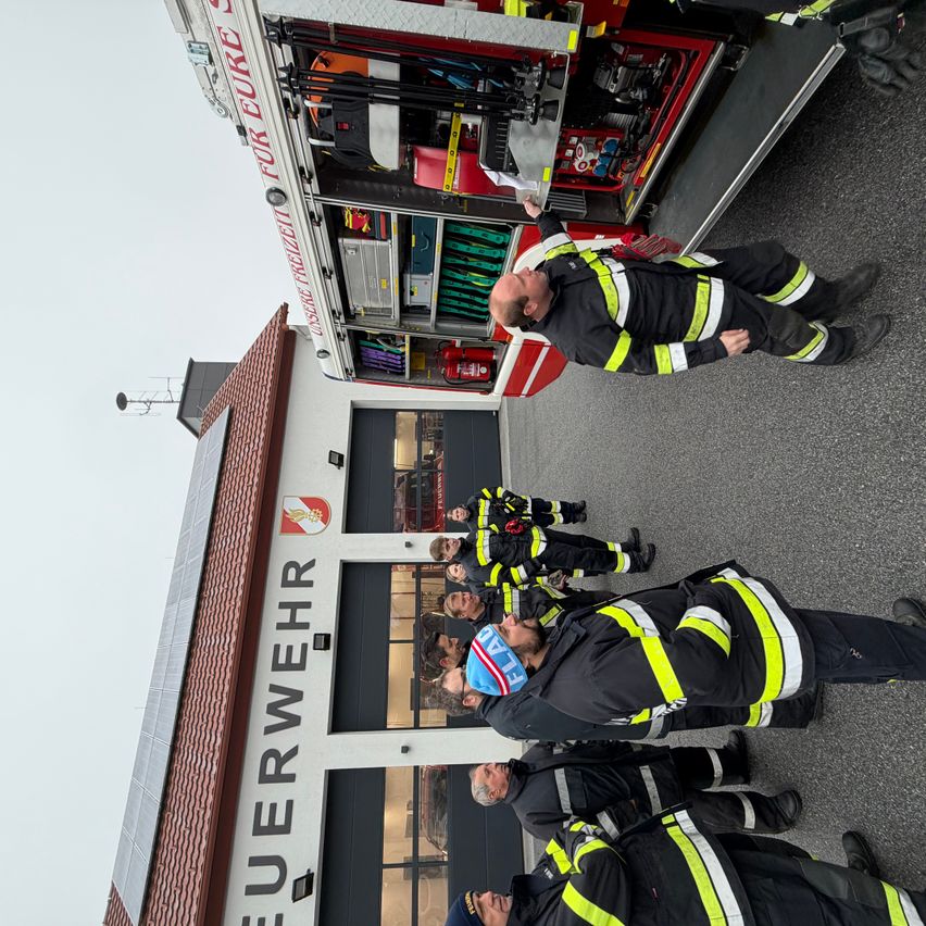 Feuerwehrleute stehen vor einer Feuerwache, ein roter Feuerwehrwagen mit den Worten 'Freiwillige Feuerwehr' ist sichtbar. Das Gebäude hat ein braunes Dach und Glasfenster.