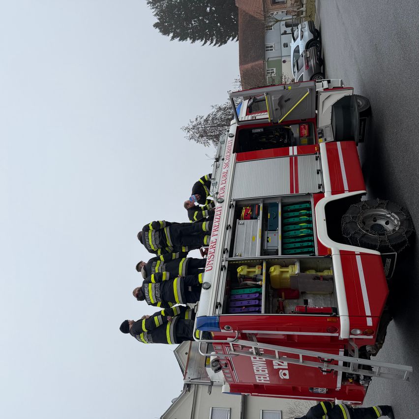 Ein Feuerwehrauto steht mit offenen Hecktüren auf einer Straße. Mehrere Feuerwehrleute stehen auf dem Lkw, und einer klettert die Leiter hoch.