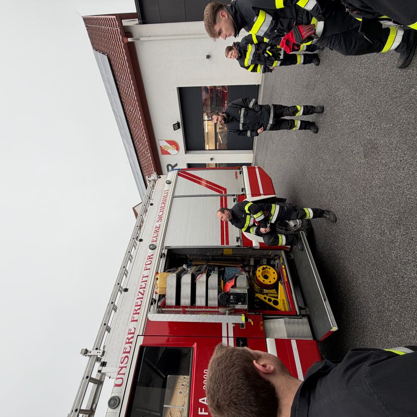Eine Gruppe von Feuerwehrleuten versammelt sich um einen roten Feuerwehrwagen mit offenen Türen. Sie bereiten sich auf eine Übung vor. Eine Leiter ist vom Wagen ausgefahren.