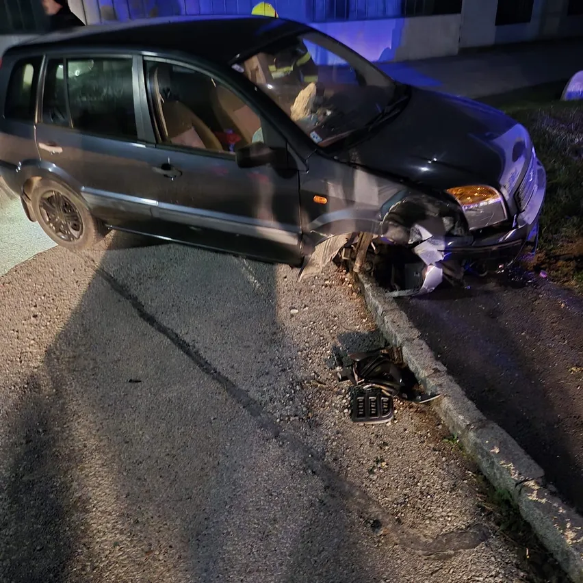 Ein dunkles Auto hat mit einem Bordstein kollidiert und hat Frontschaden erlitten. Ein Feuerwehrmann ist in der Nähe.