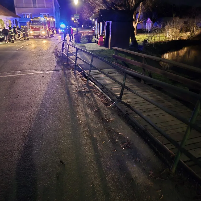Feuerwehrleute sind nachts auf der Straße, mit einem Feuerwehrwagen und eingeschalteten Lichtern. Eine kleine Brücke ist sichtbar, und eine Person steht in der Nähe eines Baumes.