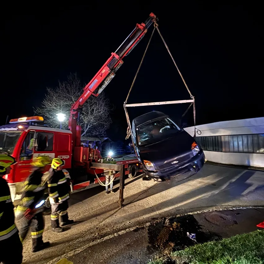 Ein Auto wird nachts von einem Kran angehoben. Drei Feuerwehrleute beobachten. Ein Mann ist im Auto. Ein Gebäude steht hinter dem Auto.