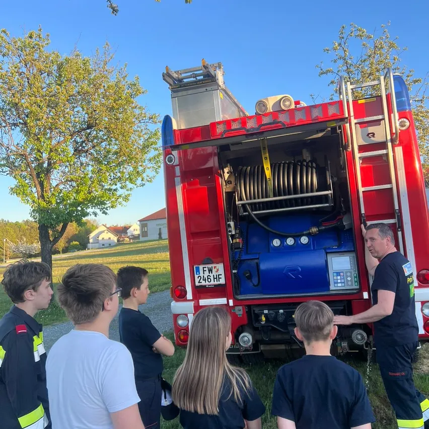 Ein Mann in Uniform zeigt Kindern das Innere eines Feuerwehrwagens. Sie stehen vor einem Baum.