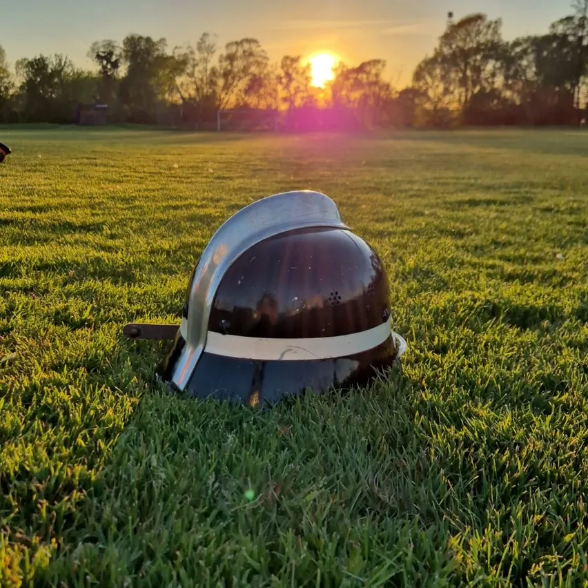 Ein Polizeihelm liegt auf einem Grasfeld mit einem Sonnenuntergang im Hintergrund. Der Himmel ist eine Mischung aus Orange und Pink.
