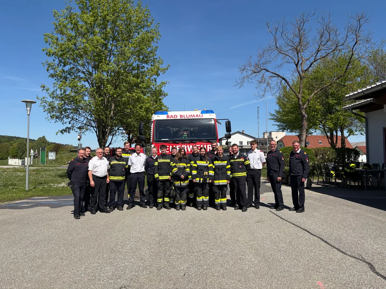 Eine Gruppe von Feuerwehrleuten posiert für ein Foto vor einem Feuerwehrwagen. Sie tragen Uniformen und Helme. Im Hintergrund sind Bäume und Gebäude sichtbar.