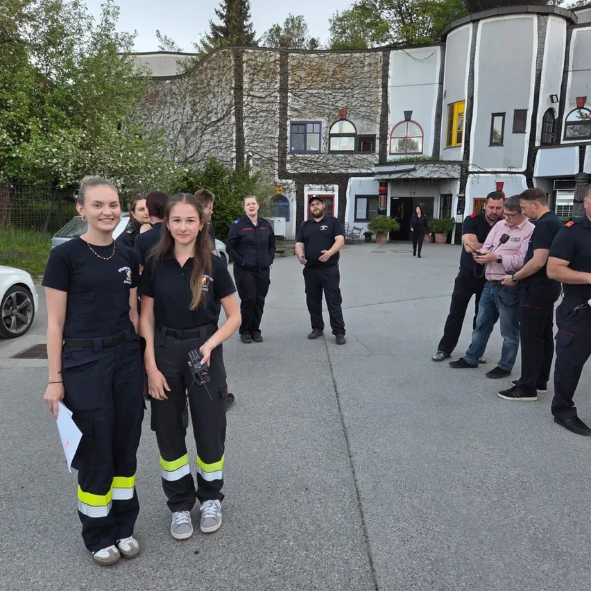 Zwei Frauen in Uniform stehen vor einem Gebäude mit einer Gruppe von Menschen im Hintergrund. Eine Frau hält ein Walkie-Talkie.