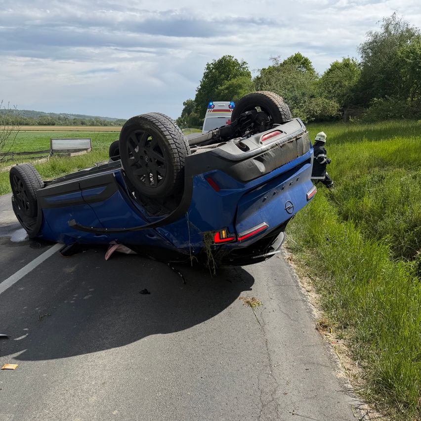 Ein blaues Auto liegt auf der Straße, ein Rad ist noch angebracht. Ein Krankenwagen ist in der Nähe, und eine Person in Schutzkleidung steht neben dem Auto.